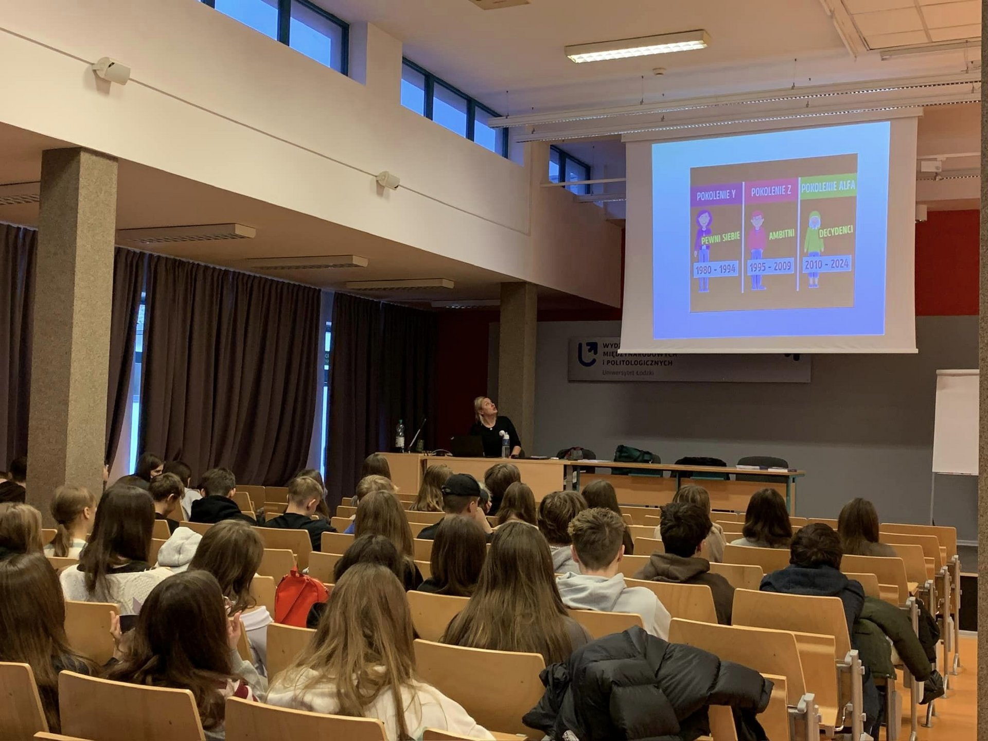 The photo shows a group of people in a lecture hall. There is a projector in the room with a multimedia presentation. A lecturer is standing at the projector.