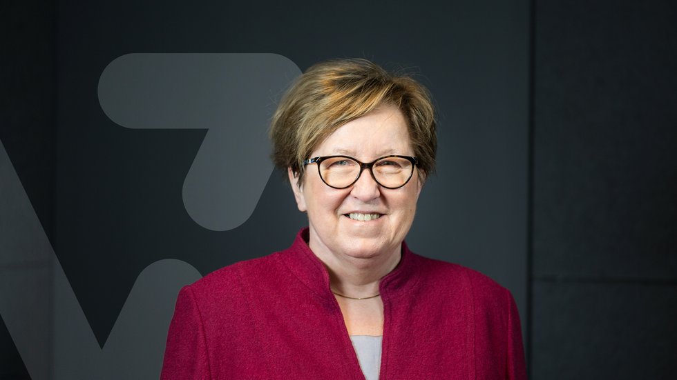 Professor Maria Bryszewska wearing a red jacket and glasses, the "V" symbol is visible in the background which is the graphic of the "Science inspires" project