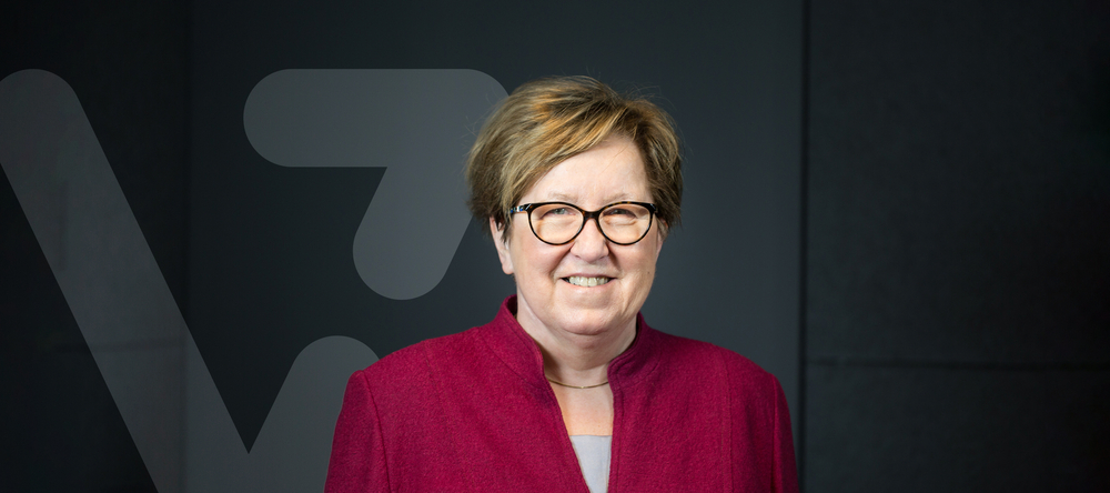 Professor Maria Bryszewska wearing a red jacket and glasses, the "V" symbol is visible in the background which is the graphic of the "Science inspires" project