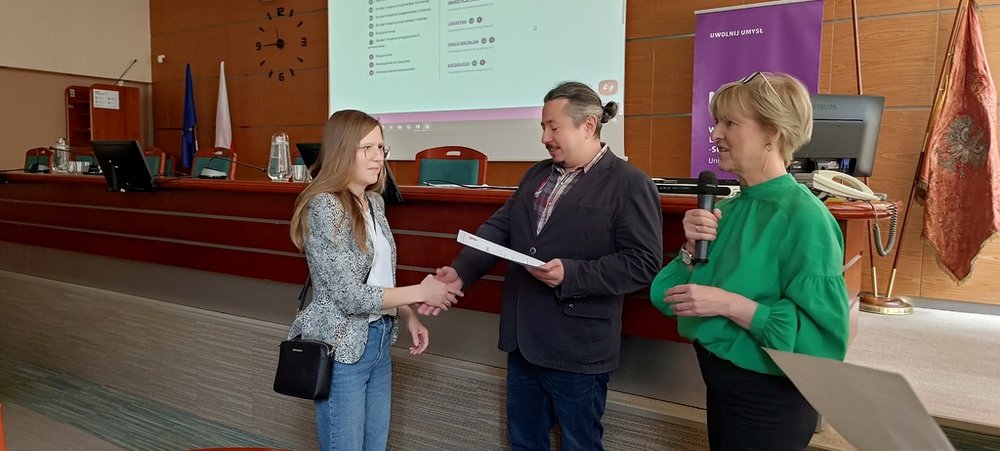 mężczyzna wręcza dziewczyninie dyplom i uściska rękę z nią, a obok nich stoi kobieta 