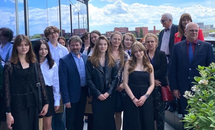 Grupa osób stojących na zewnątrz na tarasie lub balkonie budynku. Wszyscy są elegancko ubrani. Wśród grupy znajduje się zarówno młodzież, jak i dorośli. Niektóre osoby trzymają torby z logo Uniwersytetu Łódzkiego. W tle widać fragmenty miejskiego krajobrazu oraz zieleń.