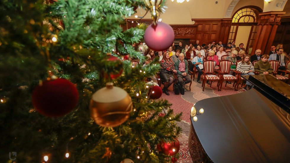 The photo shows the hall at the Biedermann’s Palace during a concert. A fragment of a Christmas tree, a piano, and a group of people sitting on chairs are visible.