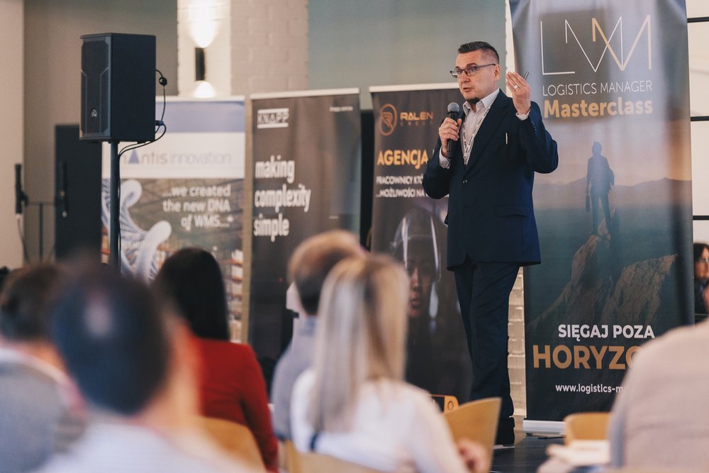 mężczyzna przemawiający przed publicznością, a za nim standy