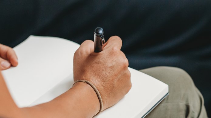 Osoba pisząca długopisem w notesie; widoczna ręka trzymająca długopis nad otwartą, pustą kartką