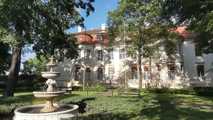 Zdjęcie przedstawia park, kamienną fontannę i w tle widoczny budynek pałacowy. Jest pałac Alfreda Biedermanna 