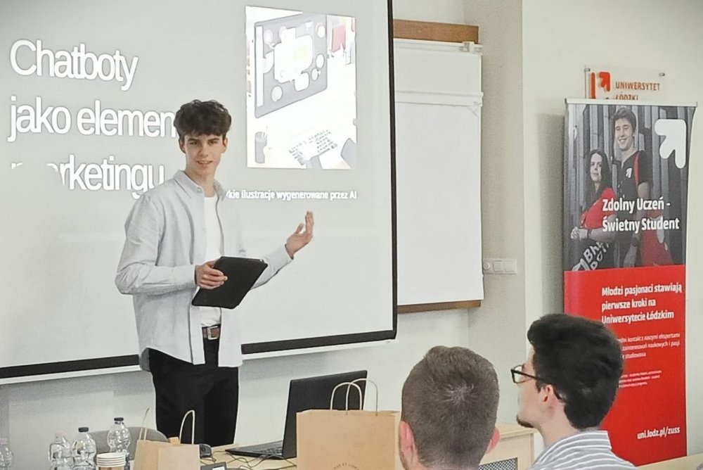 Na zdjęciu znajduje się młody mężczyzna przed ekranem projekcyjnym, na którym wyświetlany jest slajd z napisem "Chatboty jako element marketingu". Prezenter trzyma tablet w jednej ręce i wskazuje na ekran drugą ręką. W tle, obok ekranu, znajduje się baner z napisem „Zdolny uczeń-świetny student”.