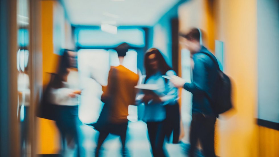 [Translate to English:] Grupa kilku osób w ruchu w korytarzu; jedna osoba trzyma dokumenty, inna zagląda w telefon. Wnętrze jest oświetlone światłem dziennym i sztucznym, występują ciepłe i chłodne barwy. Postacie są nieostre, scena sugeruje swobodną, codzienną sytuację.