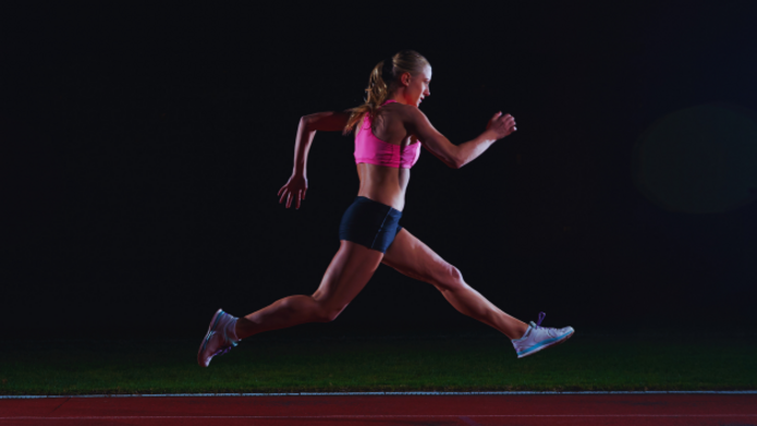 Na ciemnym tle widać młodą sportsmenkę w różowym topie i czarnych spodenkach, która dynamicznie biegnie po bieżni lekkoatletycznej. Jej sylwetka uchwycona jest w locie, co podkreśla siłę i energię ruchu.