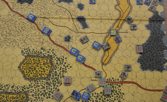 Mapa strategicznej gry planszowej z siatką heksagonalną, przedstawiająca teren z lasami, drogami i wzniesieniami. Na mapie widoczne są oznaczone miejscowości, m.in. „Starawioska” i „Tabalice”. Rozmieszczone są kolorowe żetony jednostek z literami i symbolami, reprezentujące różne frakcje wojskowe — m.in. niebieskie i żółte z oznaczeniami „C” i „P” oraz szare i czerwone z „U” i „D1”. Układ sugeruje rozgrywkę symulującą działania wojenne.