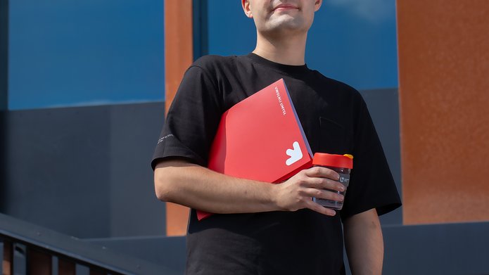 Student w okularach przeciwsłonecznych stojący z czerwoną teczką i kubkiem