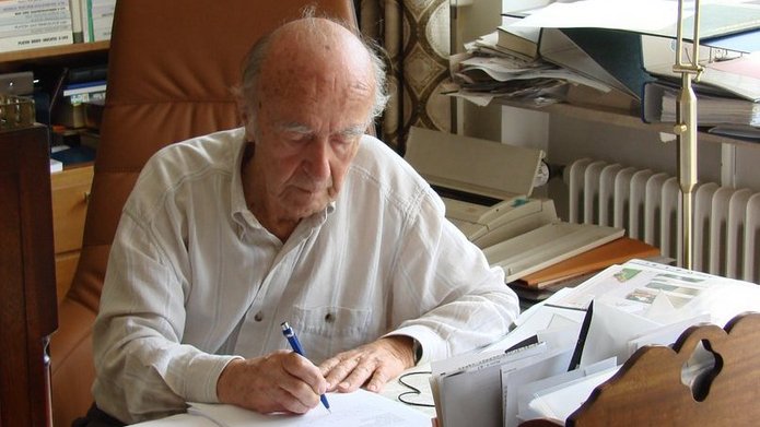  Karl Dedecius sitting at a desk with a large number of papers, books and notes, writing with a pen on a piece of paper