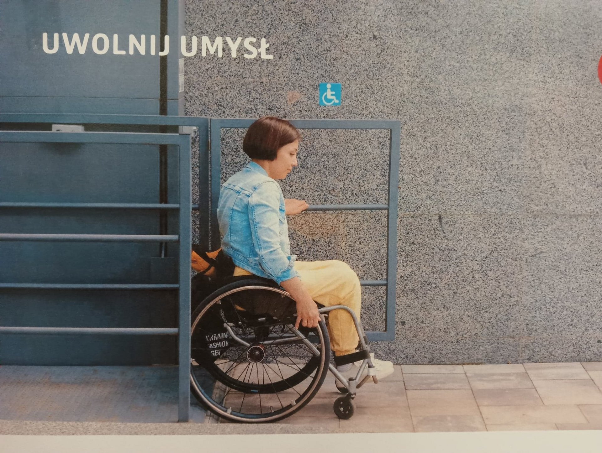 Delikatnie uśmiechająca się Studentka z niepełnosprawnością ruchową siedzi na wózku. W tle napis "Uwolnij Umysł"