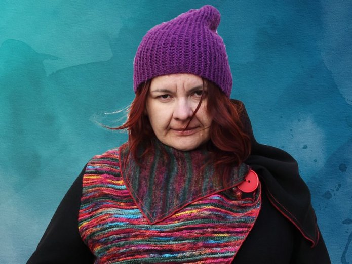 A portrait photo of the winner wearing a coat with colourful parts and a violet hat which compliments her red hair