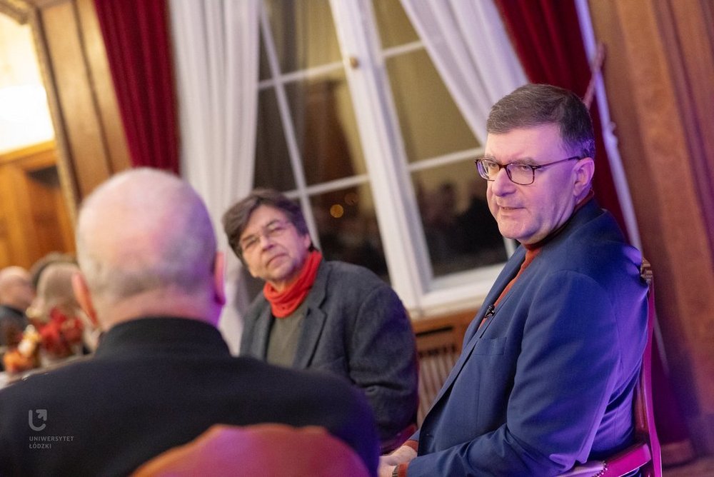 Na zdjęciu widać trzech mężczyzn. Mężczyźni siedzą przy stoliku, rozmawiają ze sobą. Widzimy twarze dwóch, jeden siedzi do nas tyłem. Są w sali, która wygląda jak sala balowa. 