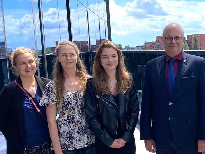 Cztery osoby stojące na zewnątrz, na tle budynku z przeszklonymi ścianami. Dwie kobiety po lewej stronie mają blond włosy i są ubrane w eleganckie stroje. Trzecia kobieta, stojąca bliżej prawej strony, ma brązowe włosy i jest ubrana w czarną skórzaną kurtkę. Mężczyzna na skrajnej prawej stronie ma na sobie ciemny garnitur z czerwoną koszulą i krawatem. W tle widać niebo z chmurami oraz fragmenty miejskiego krajobrazu z budynkami. 