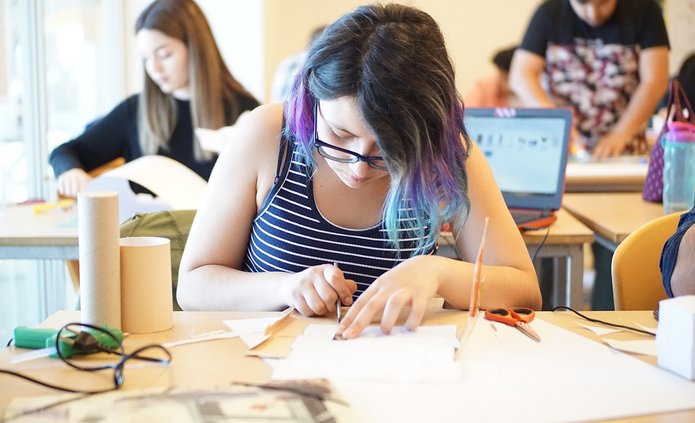 Na obrazku widzimy grupę młodych osób pracujących przy stolikach. Na pierwszym planie znajduje się dziewczyna z długimi włosami o kolorze w tonach fioletu i niebieskiego. Ma na sobie bluzkę w poziome, czarno-białe paski. Skupia się na swojej pracy, korzystając z narzędzi do rękodzieła lub projektowania — używa noża lub nożyczek do precyzyjnego cięcia papieru. Obok niej na stoliku znajdują się różne przedmioty: tubki kartonowe, nożyczki, laptop, i inne akcesoria plastyczne. W tle widać inne osoby, które również są zaangażowane w różne zadania, prawdopodobnie związane z projektem lub zajęciami manualnymi. Atmosfera jest typowa dla pracowni lub sali warsztatowej, gdzie studenci skupiają się na kreatywnej pracy.