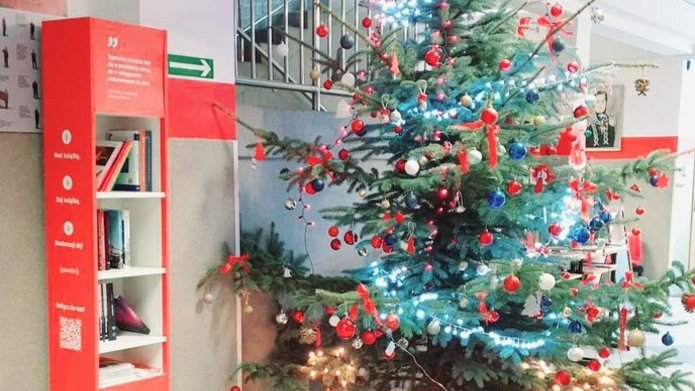 Zdjęcie przedstawia korytarz w szkole. Stoi choinka. Obok niej stoi czerwony regał z książkami. 