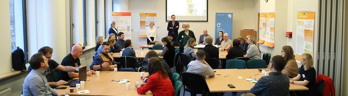 grupa osób siedzi w sali, a dwóch prowadzących omawia temat