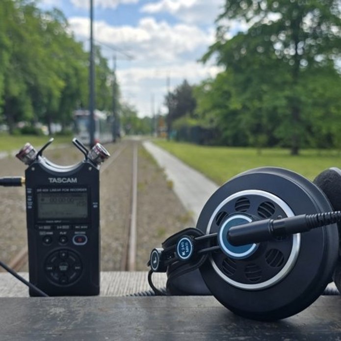 Na zdjęciu widać urządzenie nagrywające typu dyktafon w kolorze czarnym, po prawej stronie leżą słuchawki. W drugim planie o oddali widać fragment parku, zielone drzewa, fragment niebieskiego nieba.
