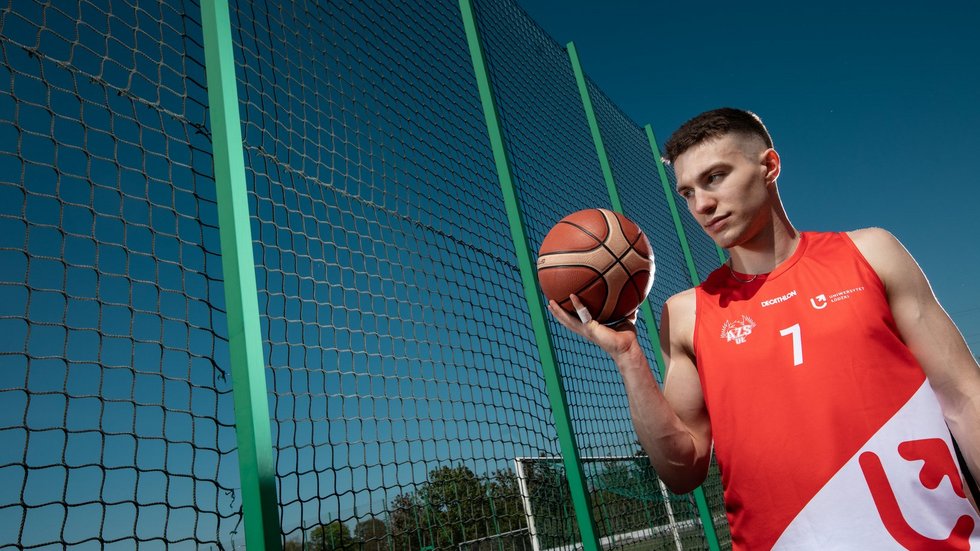 Wysoki chłopak z piłka do koszykówki z ręku stoi na biosku do koszykówki w stroju sportowym.