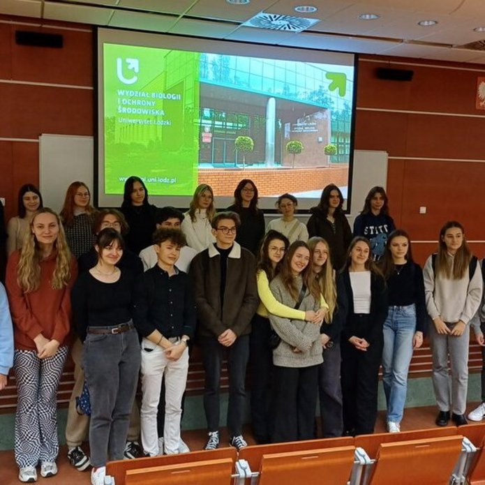 Grupa nastolatków stoi w auli na tle ekranu z prezentacją multimedialną 