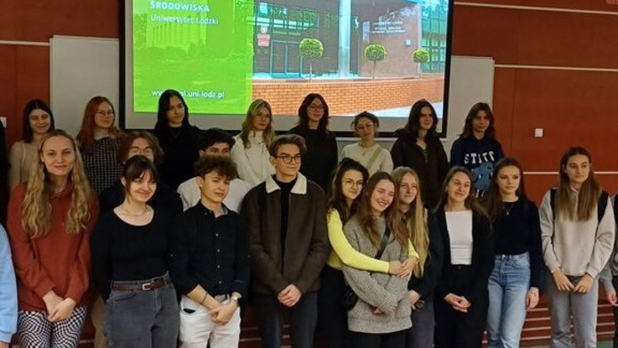 Grupa nastolatków stoi w auli na tle ekranu z prezentacją multimedialną 