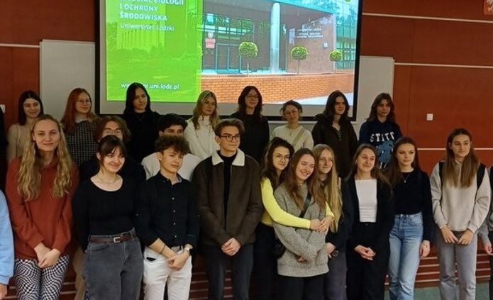 Grupa nastolatków stoi w auli na tle ekranu z prezentacją multimedialną 