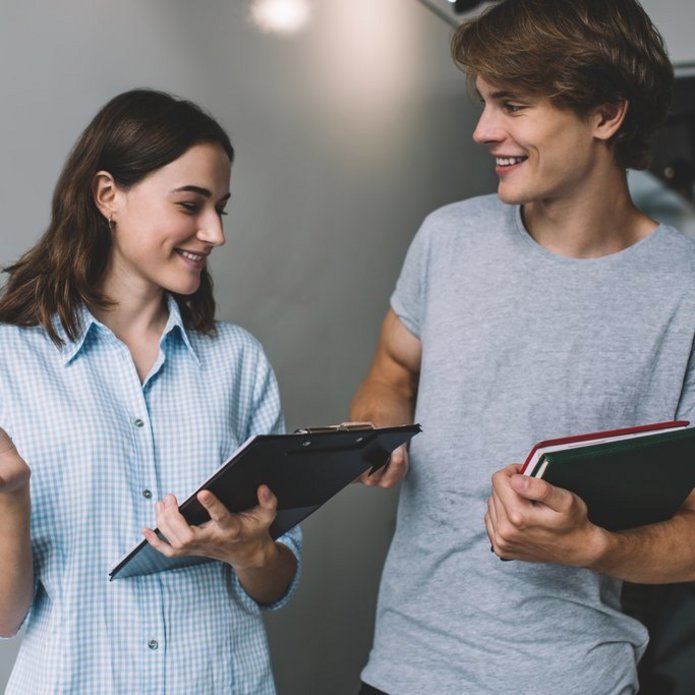 Dwoje uśmiechniętych studentów stoi obok siebie — jeden trzyma notesy, drugi podkładkę na dokumenty.