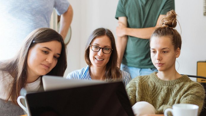 grupa uśmiechniętych młodych ludzi siedzi w pokoju, patrzą na ekran laptopa 