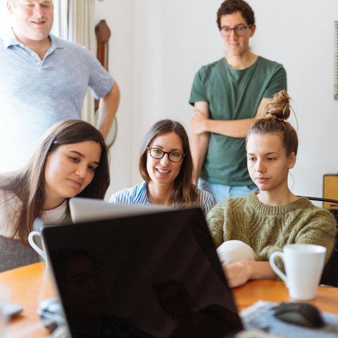 group of smiling young people sitting in a room, looking at a laptop screen