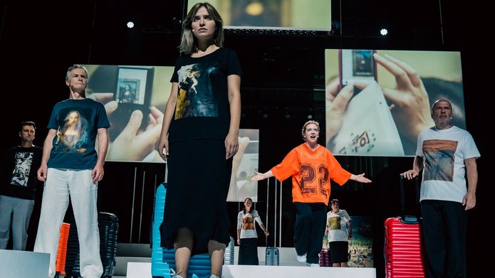 Na zdjęciu widoczna jest grupa osób stojących na scenie. Osoby są ubrane jak turyści, mają walizki. Za nimi wiszą różnej wielkości plansze na których są zdjęcia z różnych turystycznych miejsc.