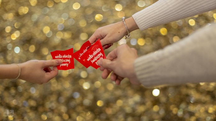 3 ręce trzymające czerwone serduszka z napisem Wielka Orkiestra świątecznej pomocy na tle złotych monet