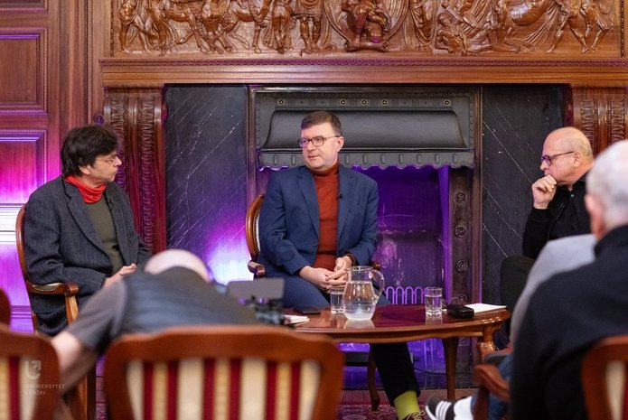 Na zdjęciu widać trzech mężczyzn. Mężczyźni siedzą przy stoliku, rozmawiają ze sobą. Są w sali, która wygląda jak sala balowa. Za nimi widać kominek, w którym nie pali się ogień. 