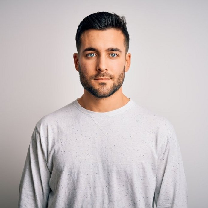 Młody mężczyzna o ciemnych, krótko przystrzyżonych włosach i gęstej, przyciętej brodzie, ubrany w jasnoszary sweter, stoi przodem do aparatu na jednolitym, jasnym tle, patrząc spokojnie w obiektyw.