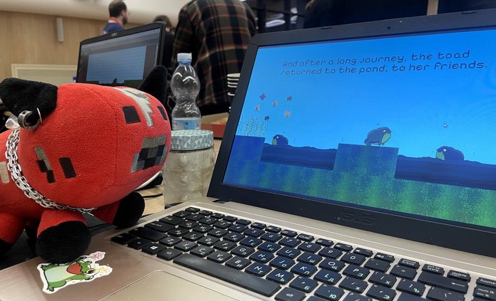 Na zdjęciu widać otwarty laptop stojący na stole podczas wydarzenia. Na ekranie wyświetlana jest gra komputerowa w stylu pikselowym z żabą na tle nieba i wody oraz tekstem końcowym gry. Obok laptopa leży czerwono-czarna pluszowa maskotka w kształcie zwierzątka z kolczykiem i łańcuszkiem. Na stole znajdują się także butelka wody i słoik. W tle widać grupę osób stojących w sali, co sugeruje, że zdjęcie zostało wykonane podczas maratonu tworzenia gier.