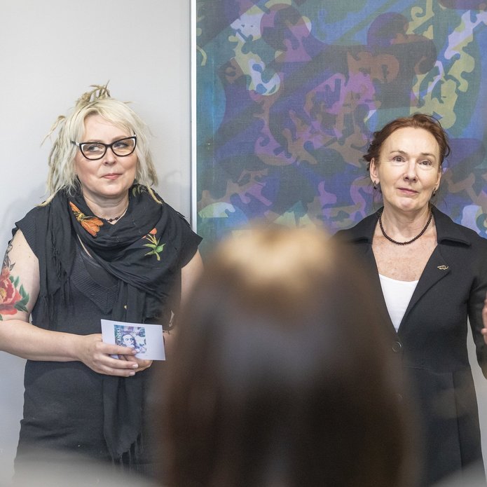 Na zdjęciu widoczne są dwie kobiety. Stoją twarzami do nas. Za nimi widczone są wiszące na ścianie obrazy. Kobiety widzimy od pasa w górę. 