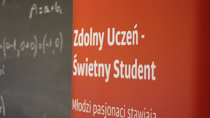Na zdjęciu jest czerwony baner z białymi napisami: Zdolny uczeń - świetny student. W tle jest czarna tablica z zapisanym działaniem matematycznym