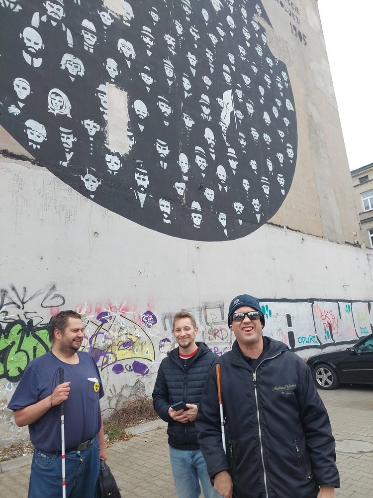 three smiling people standing in front of one of the Lodz murals, two of them have white sticks with them