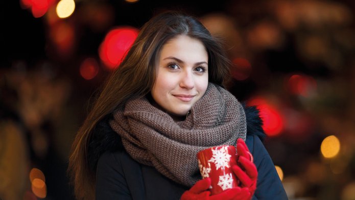 uśmiechnięta kobieta z długimi włosami ubrana w kurtkę, brązowy szalik i czerwone rękawiczki, trzymająca czerwony kubek z białymi płatkami śniegu, w tle świąteczne światła