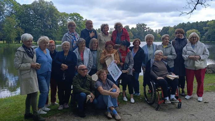 Na zdjęciu widać grupę około 20 osób. To mężczyźni i kobiety w dojrzałym wieku, pozują do zdjęcia grupowego. Stoją w plenerze. Jest to pora letnia.