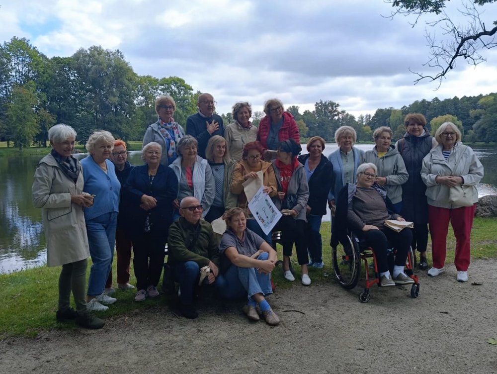 The photo shows a group of about 20 people. They are mature men and women, posing for a group photo. They are standing outdoors. It's summer.