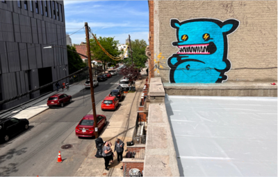 po prawej  stronie fikcyjna niebieska postać na murze, street art, po lewej ulica, samochody