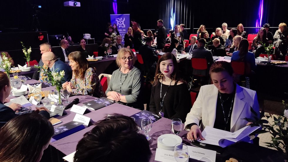 Na zdjęciu widoczne są osoby siedzące za stołem. Jest to spora grupa. Osoby debatują w sali, która wygląda jak sala teatralna. Jest dość ciemno, a oświetlenie jest teatralne, z fioletowym odcieniem. 