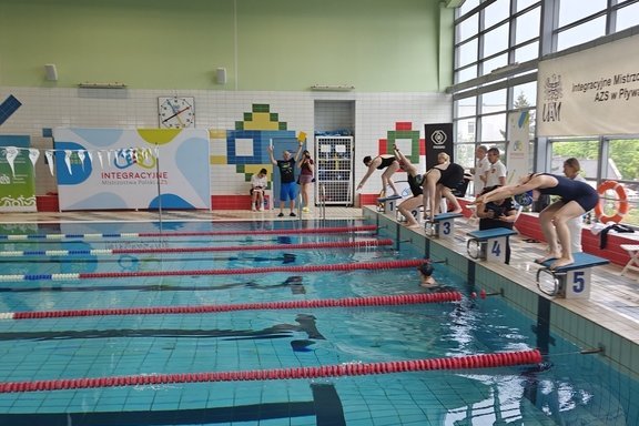 Wnętrze krytej pływalni podczas zawodów pływackich. Kilku zawodników w strojach kąpielowych stoi na słupkach startowych, przygotowując się do skoku do wody. Jeden z zawodników jest już w trakcie skoku. 