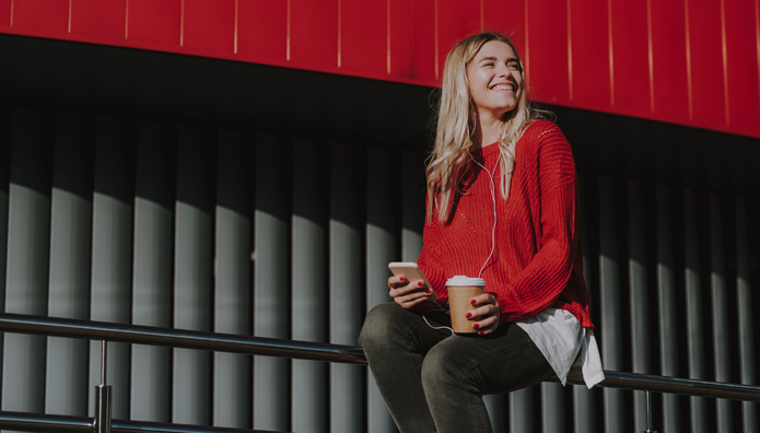 Na zdjęciu widoczna jest młoda kobieta siedząca na metalowej barierce. Ma na sobie czerwony sweter, ciemne spodnie i białą koszulę wystającą spod swetra. W jednej dłoni trzyma smartfon, w drugiej papierowy kubek z kawą; w uszach ma słuchawki. Kobieta uśmiecha się i patrzy w bok. Tło stanowi nowoczesna architektura z pionowymi, ciemnymi panelami oraz intensywnie czerwonym elementem konstrukcyjnym u góry kadru.