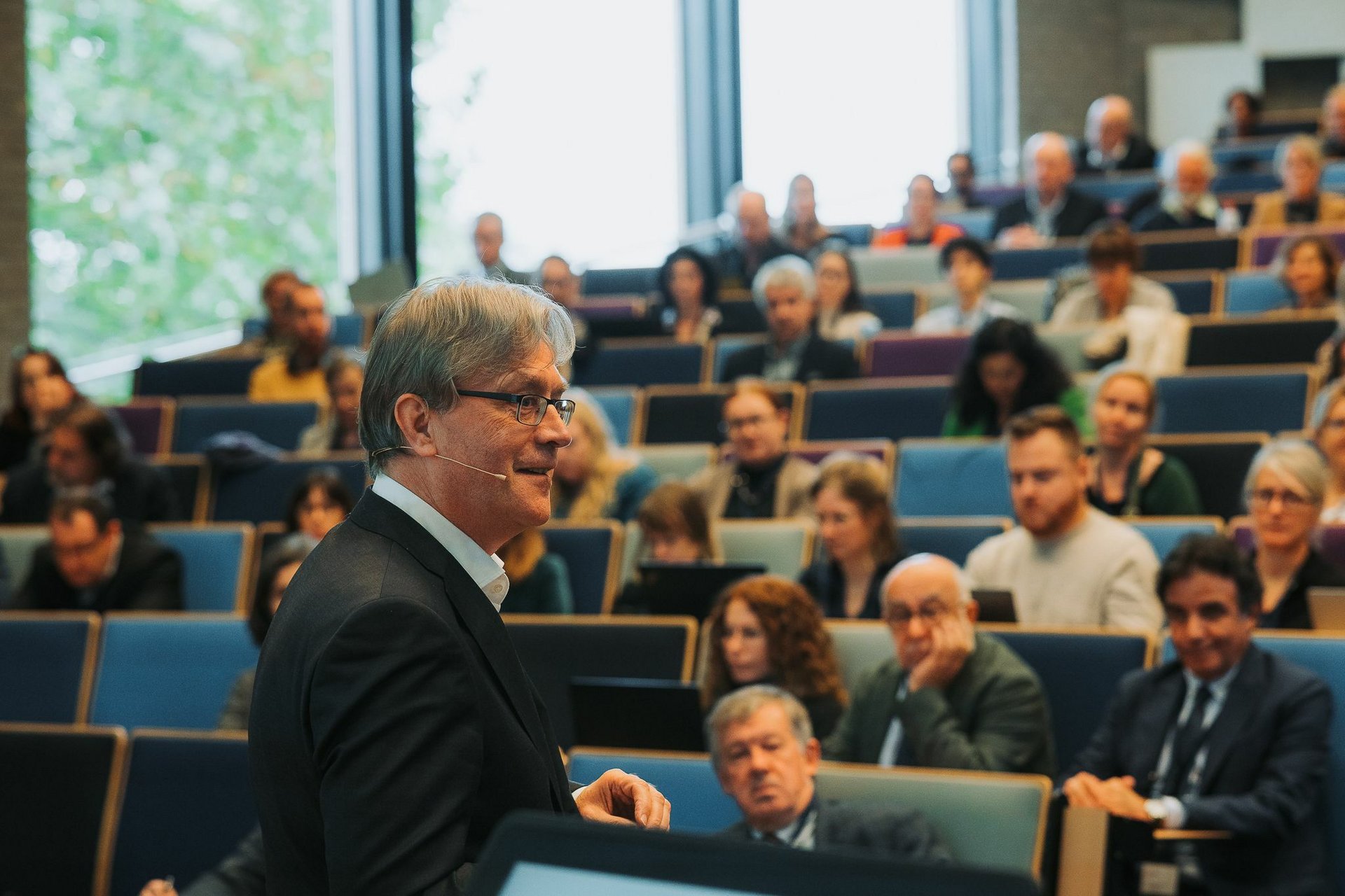 A man looking right and talking to the microphone; in the background the audience at the university hall