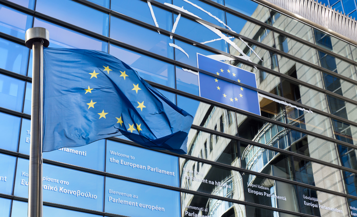 Na pierwszym planie powiewa flaga Unii Europejskiej na maszcie. W tle widoczna jest nowoczesna, przeszklona fasada budynku Parlamentu Europejskiego z napisem powitalnym w różnych językach oraz emblematem UE.