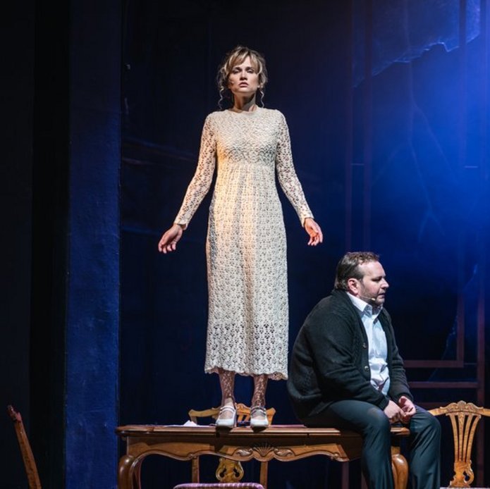 Fotos ze spektaklu. Kobieta stoi na stole, obok siedzi mężczyzna.