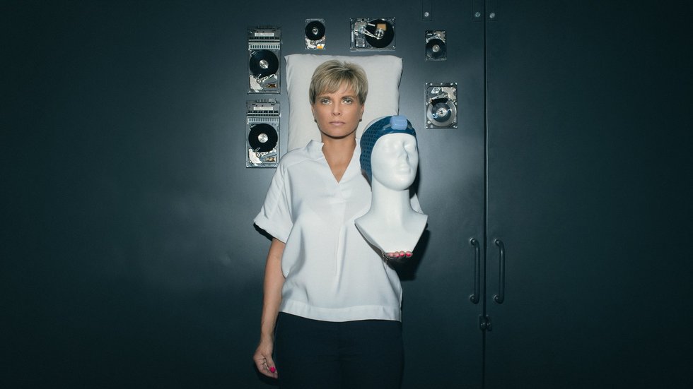 Prof. Renata Bocian holding a mannequin wearing a Vguard band, with a pillow and floating computer hard drives in the background