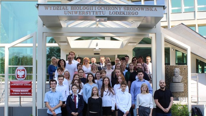 Duża grupa osób stojących przed budynkiem oznaczonym napisem "Wydział Biologii i Ochrony Środowiska Uniwersytetu Łódzkiego". Grupa składa się z młodych ludzi i dorosłych. Wszyscy są elegancko ubrani, niektórzy w białe koszule, inni w ciemne marynarki.W tle widać przeszklone drzwi budynku oraz tabliczkę z godłem Polski po lewej stronie.